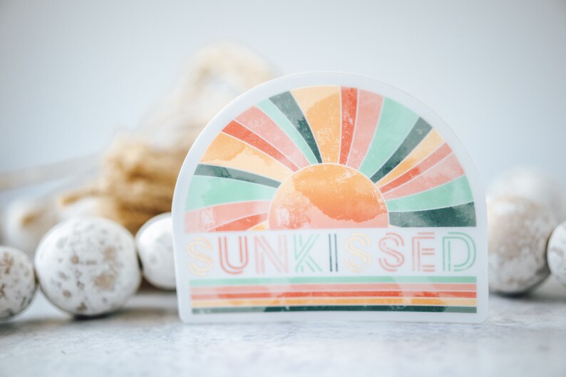 May include: A white sticker with a colourful sunburst design and the text "SUNKISSED" in a retro font. The sticker is half-circle shaped and has a watercolour effect.