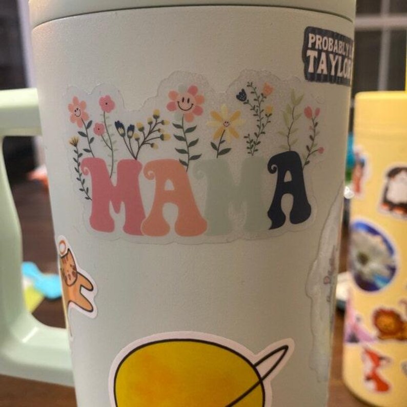 Pode incluir: Uma garrafa de &aacute;gua reutiliz&aacute;vel azul claro com um adesivo branco que diz "MAMA" em uma fonte retr&ocirc; com letras rosa, laranja e azul. O adesivo &eacute; cercado por um design floral com flores rosa, amarelas e verdes. H&aacute; um pequeno adesivo de um gato na garrafa.