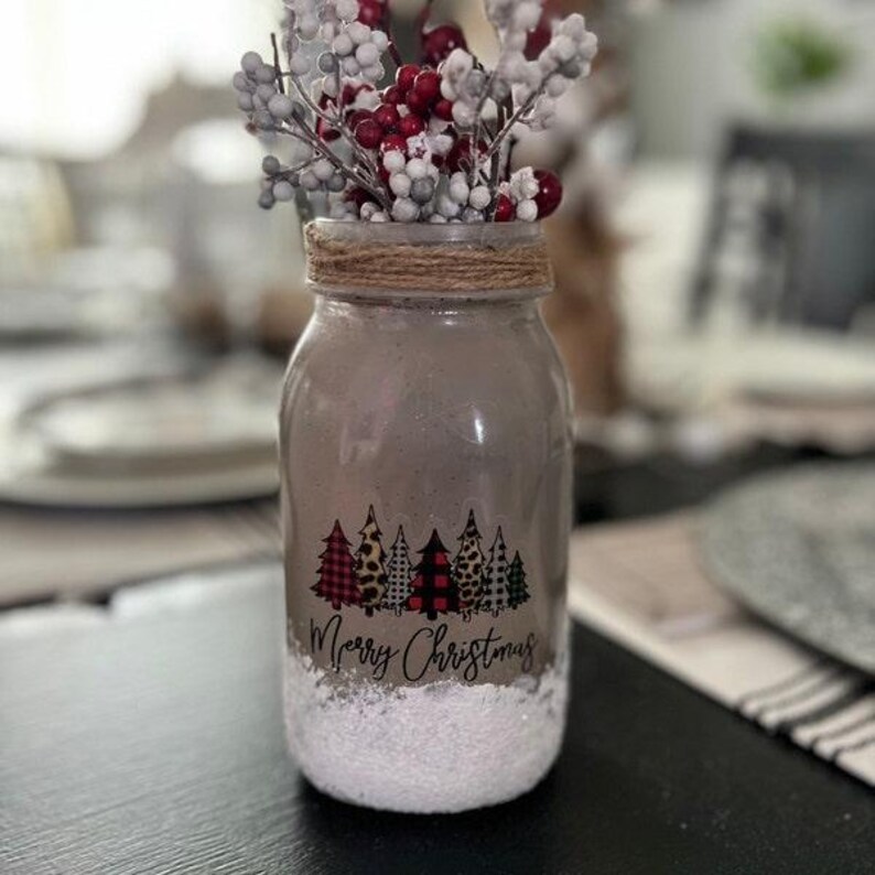 Puede incluir: Un tarro de cristal decorado con una escena de invierno. El tarro est&aacute; lleno de nieve artificial y tiene una pegatina con las palabras "Feliz Navidad" y una fila de &aacute;rboles de Navidad con patrones de cuadros, leopardo y otros. El tarro est&aacute; coronado con una rama de bayas y vegetaci&oacute;n artificiales.
