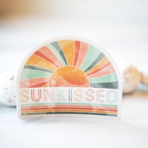 May include: A white sticker featuring a colourful retro sunburst design and the word "SUNKISSED" in a stylised font.
