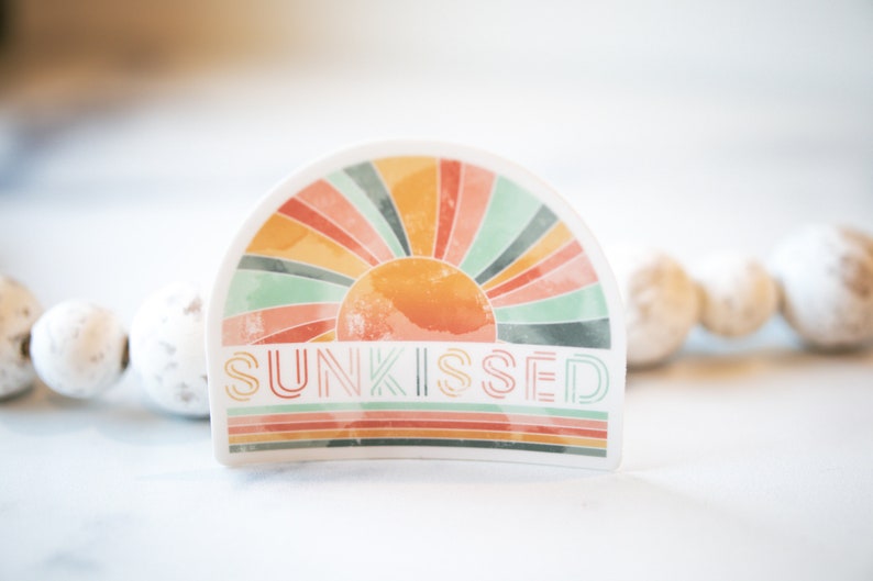 May include: A white sticker featuring a colourful retro sunburst design and the word "SUNKISSED" in a stylised font.