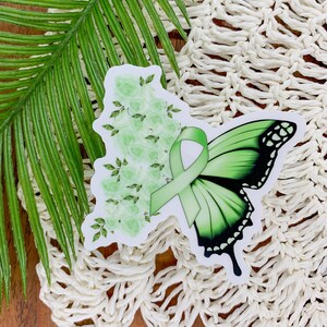 May include: Green butterfly sticker with a green ribbon and white flowers. The sticker is on a white crocheted surface.