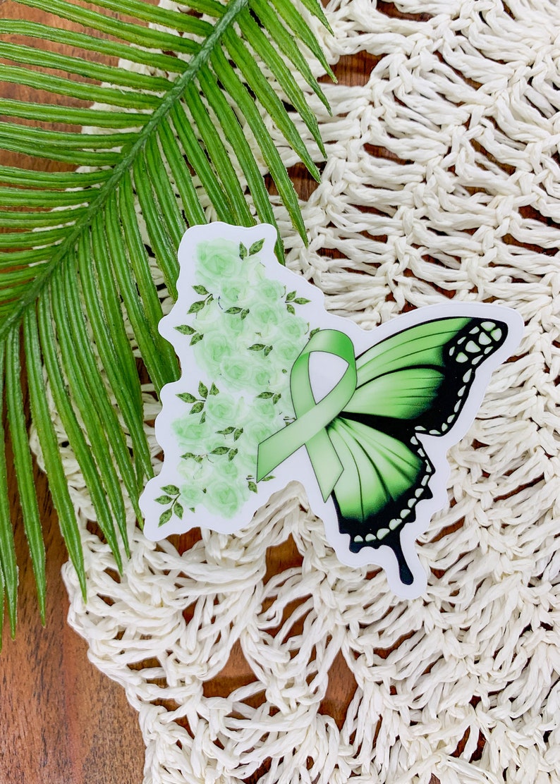 May include: Green butterfly sticker with a green ribbon and white flowers. The sticker is on a white crocheted surface.