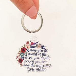 May include: A clear acrylic keychain with a floral design and the text "may you be proud of the work you do, the person you are and the difference you make".