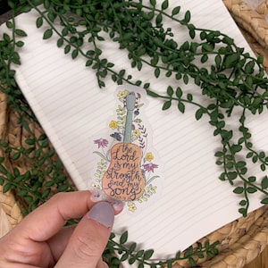 Puede incluir: Una pegatina transparente con un dise&ntilde;o floral alrededor de una guitarra. La guitarra es naranja y tiene el texto "The Lord is my strength and my song" con "Psalm 118:14" debajo.