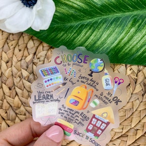 May include: A gold sticker with a back to school theme. It features illustrations of school supplies, a backpack, a school building, and a globe. The sticker has the text "Choose to be positive", "Every day is a new adventure", "You can learn something new everyday", "Be kind", "It's OK to make mistakes", "The more you read the more things you will know the more you learn on the places you'll go", and "Back to school".
