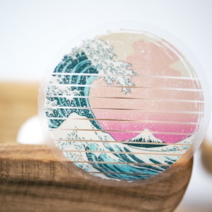 May include: A clear round sticker with a graphic of a blue and white wave crashing over a pink and white mountain. The sticker has a grid pattern over the image.