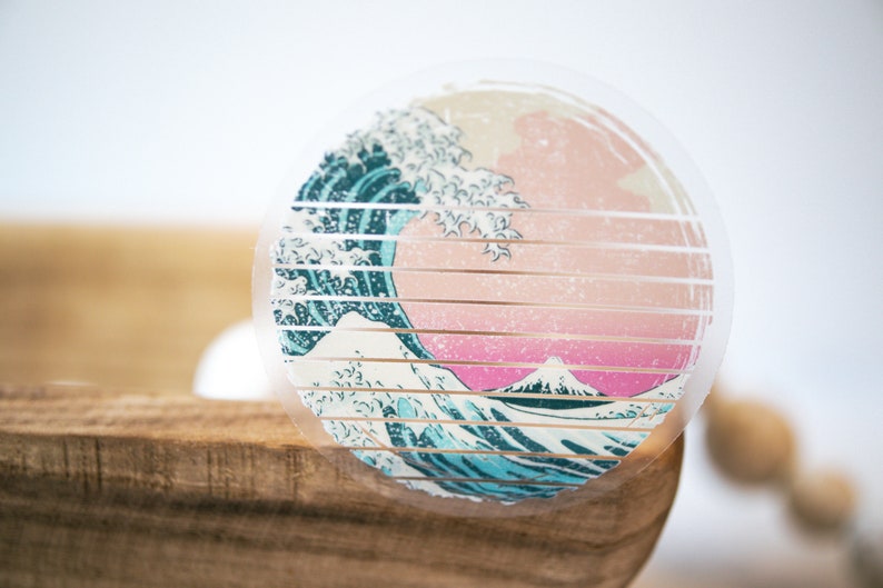 May include: A clear round sticker with a graphic of a blue and white wave crashing over a pink and white mountain. The sticker has a grid pattern over the image.