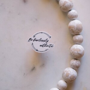May include: White round sticker with black text that reads "Be fearlessly authentic" surrounded by a black floral design. The sticker is on a white marble surface with a white beaded garland in the background.