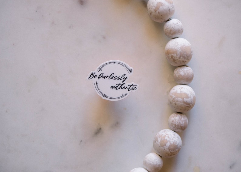 May include: White round sticker with black text that reads "Be fearlessly authentic" surrounded by a black floral design. The sticker is on a white marble surface with a white beaded garland in the background.
