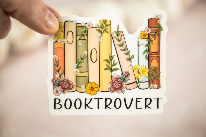 Pu&ograve; includere: Un adesivo con una pila di libri colorati con accenti floreali e il testo "BOOKTROVERT".