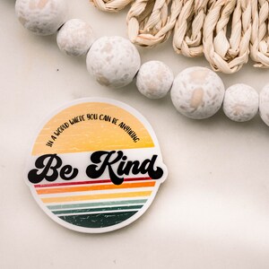 May include: A round sticker with a retro-style design featuring a yellow and orange sunset with multi-colored stripes. The text "In a world where you can be anything, Be Kind" is written in black on the sticker.