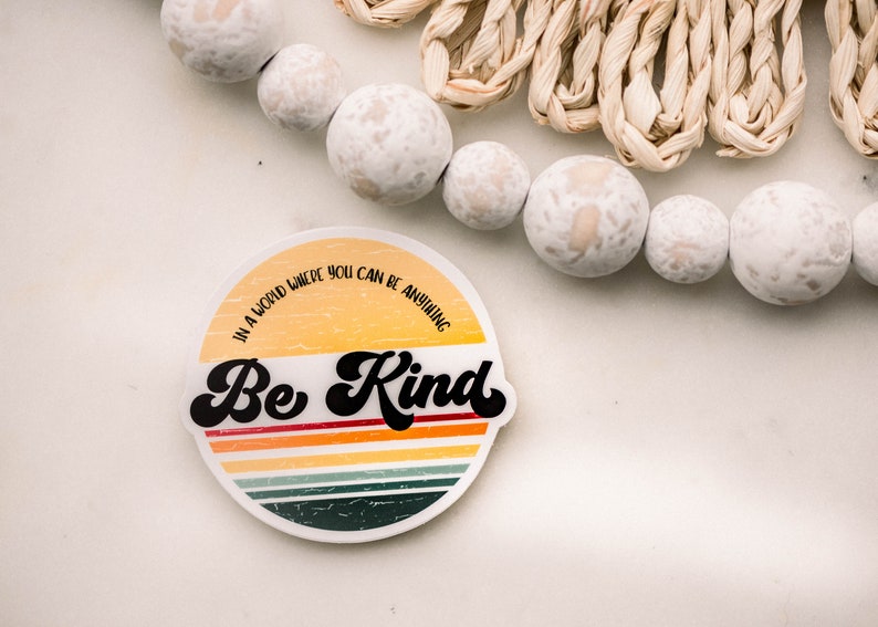 May include: A round sticker with a retro-style design featuring a yellow and orange sunset with multi-colored stripes. The text "In a world where you can be anything, Be Kind" is written in black on the sticker.