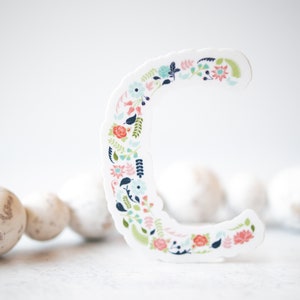 Pu&ograve; includere: Un ritaglio di carta bianca della lettera C decorato con un motivo floreale in tonalit&agrave; di rosa, blu e verde. La lettera &egrave; appoggiata su una superficie bianca con una collana di perline bianche sullo sfondo.