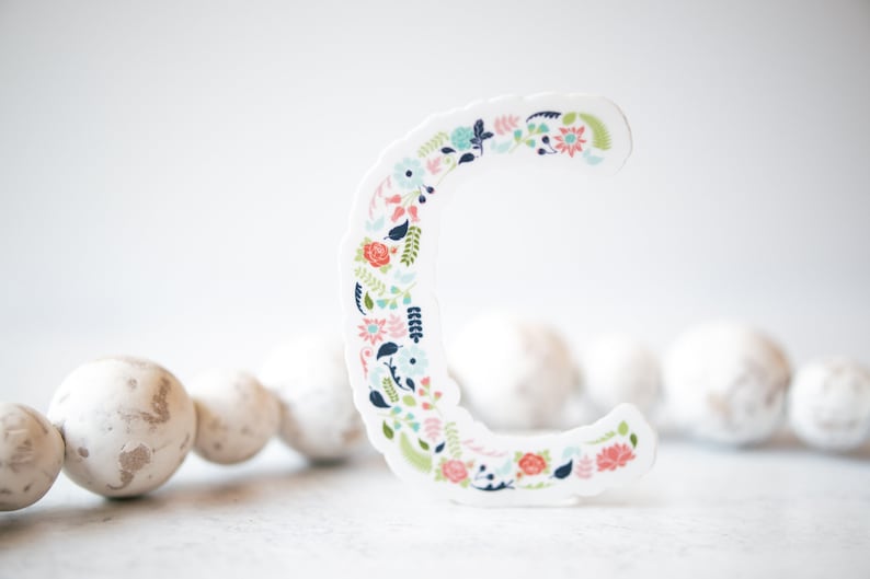 Pu&ograve; includere: Un ritaglio di carta bianca della lettera C decorato con un motivo floreale in tonalit&agrave; di rosa, blu e verde. La lettera &egrave; appoggiata su una superficie bianca con una collana di perline bianche sullo sfondo.
