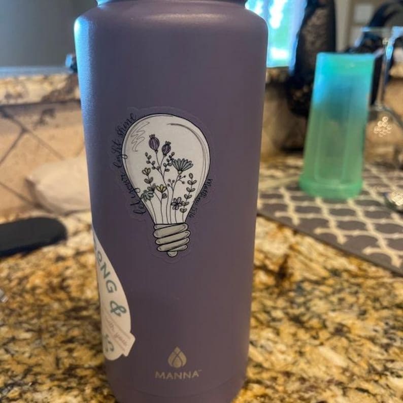 May include: Purple water bottle with a white lightbulb sticker that says "Let your light shine" with a floral design inside the lightbulb. The bottle has the word "MANNA" on the bottom.
