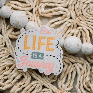 May include: A white sticker with black outlines and pink and yellow lettering that reads "Life is a Journey". The sticker is on a woven straw background.