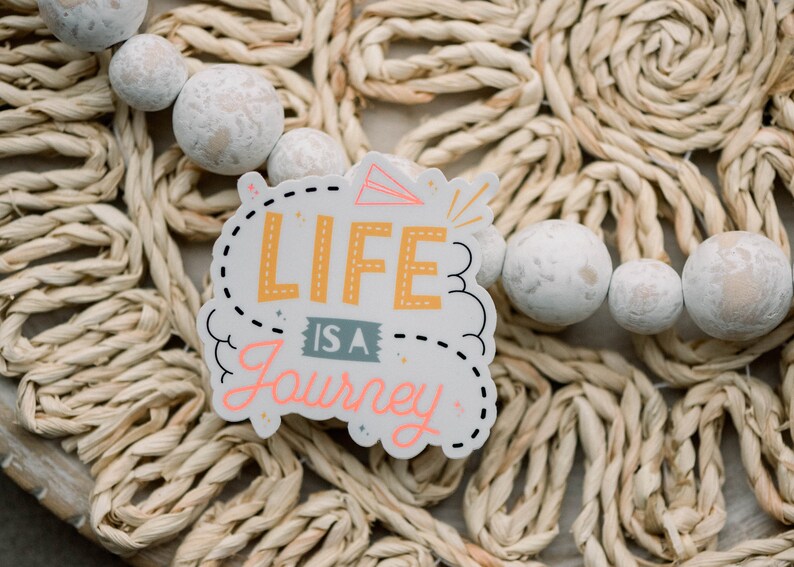 May include: A white sticker with black outlines and pink and yellow lettering that reads "Life is a Journey". The sticker is on a woven straw background.