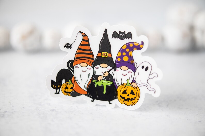 Puede incluir: Tres gnomos vestidos con trajes de Halloween, con sombreros naranja, morado y negro, sostienen una caldera y calabazas. Un gato negro y una ara&ntilde;a est&aacute;n en el fondo.