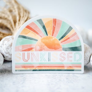 May include: A white sticker featuring a colourful rainbow sun design and the text "SUNKISSED" in a retro font.