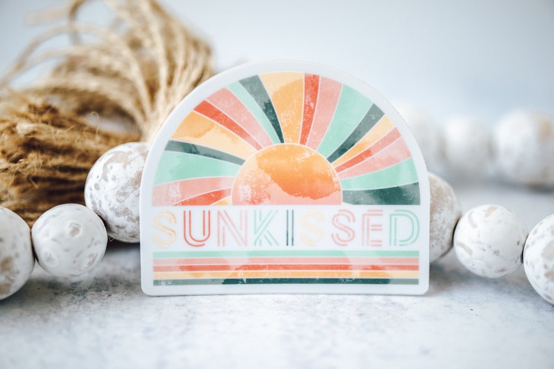 May include: A white sticker featuring a colourful rainbow sun design and the text "SUNKISSED" in a retro font.