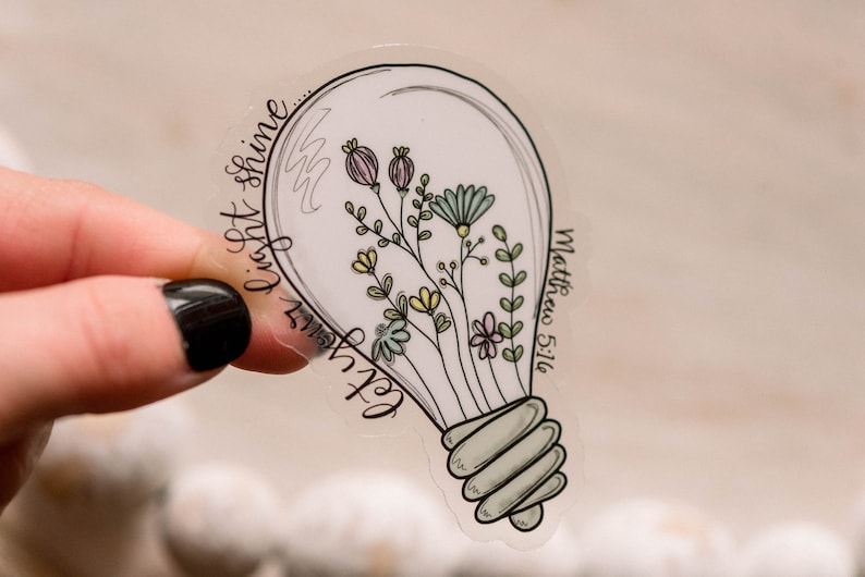 May include: A clear sticker featuring a light bulb design. Inside the bulb are colourful flowers and leaves. The text "Let your light shine" is written around the bulb and "Matthew 5:16" is written on the side.