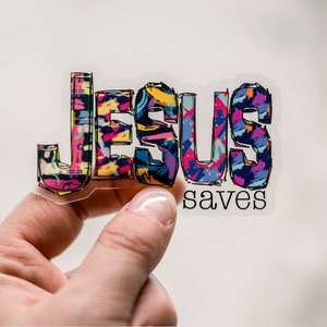 May include: A clear sticker with the word "JESUS" in colorful, patterned letters and the words "saves" in black text below.