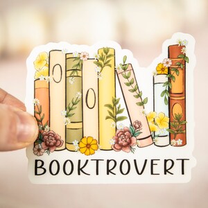 Pu&ograve; includere: Un adesivo bianco con una pila di libri colorati decorati con fiori e vegetazione. Il testo "BOOKTROVERT" &egrave; stampato sotto i libri.