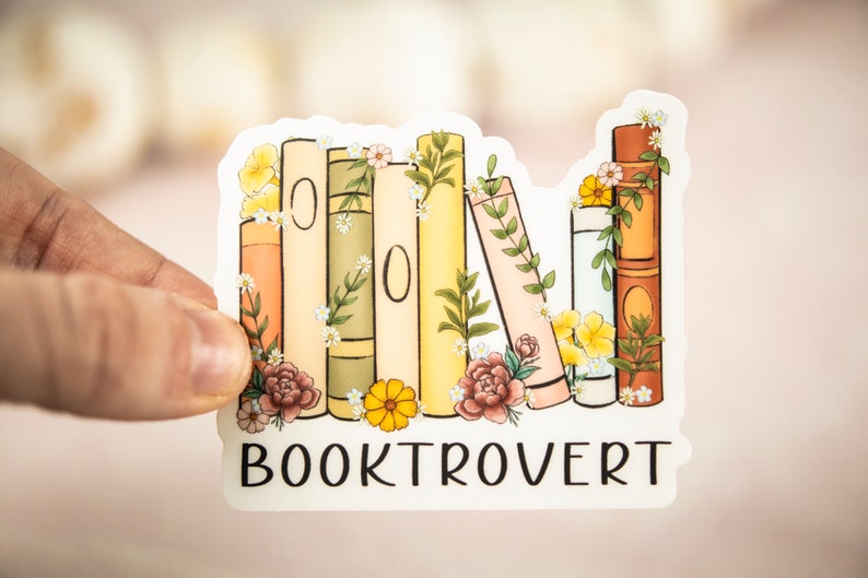 Pu&ograve; includere: Un adesivo bianco con una pila di libri colorati decorati con fiori e vegetazione. Il testo "BOOKTROVERT" &egrave; stampato sotto i libri.
