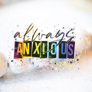 May include: A clear sticker with the words "always anxious" in black script and block letters. The word "ANXIOUS" is in a rainbow gradient. Colorful paint splatters surround the text.