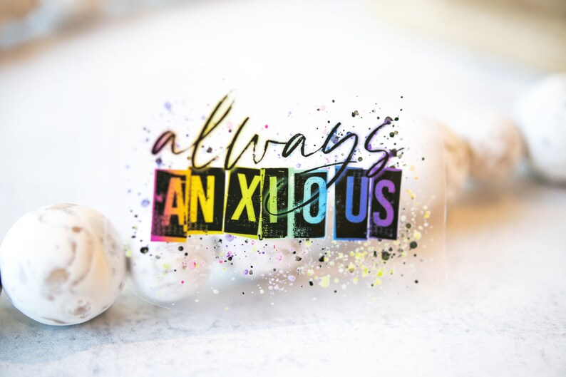 May include: A clear sticker with the words "always anxious" in black script and block letters. The word "ANXIOUS" is in a rainbow gradient. Colorful paint splatters surround the text.