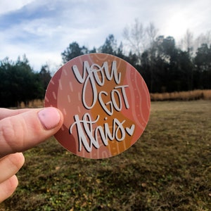 May include: A round sticker with an abstract design in shades of orange, pink, and brown. The sticker features the text "You Got This" in white with a small heart.