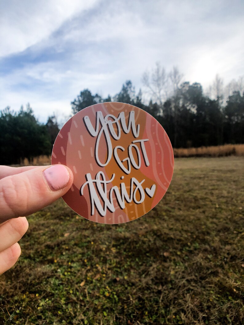 May include: A round sticker with an abstract design in shades of orange, pink, and brown. The sticker features the text "You Got This" in white with a small heart.