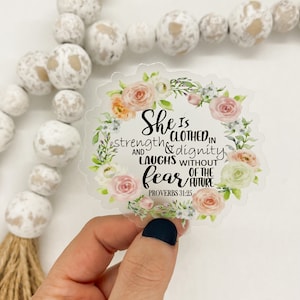 Könnte beinhalten: Ein klarer Aufkleber mit einem floralen Kranzdesign. Der Aufkleber zeigt den Text "She is clothed in strength & dignity and laughs without fear of the future. Proverbs 31:25" in schwarzer Schrift. Der Kranz enthält rosa und pfirsichfarbene Rosen.