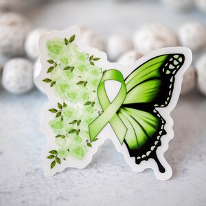 K&ouml;nnte beinhalten: Ein schmetterlingsf&ouml;rmiger Aufkleber mit einem gr&uuml;nen Band und floralem Design. Die linke Seite zeigt hellgr&uuml;ne Rosen und Bl&auml;tter, w&auml;hrend die rechte Seite einen Farbverlauf von Gr&uuml;n und Schwarz aufweist. Der Aufkleber befindet sich auf wei&szlig;em Hintergrund.