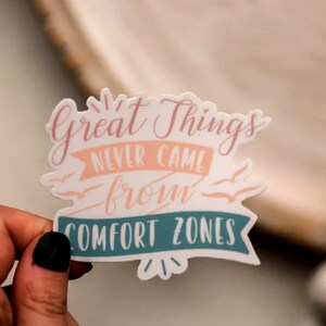 May include: A white sticker with the text "Great things never came from comfort zones" in pink and teal lettering. The sticker has a banner design with a pink background and a teal border.