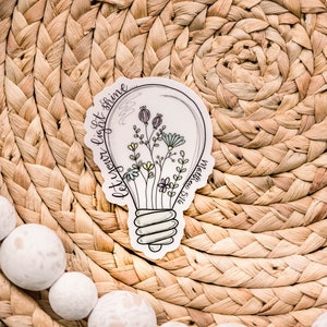 May include: A white sticker featuring a light bulb design. The light bulb contains flowers and the text "Let your light shine" surrounding it.