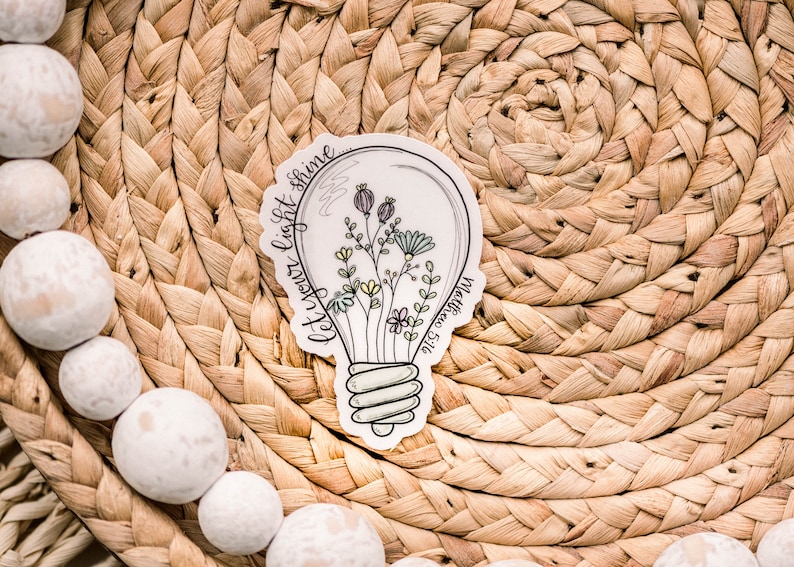 May include: A white sticker featuring a light bulb design. The light bulb contains flowers and the text "Let your light shine" surrounding it.