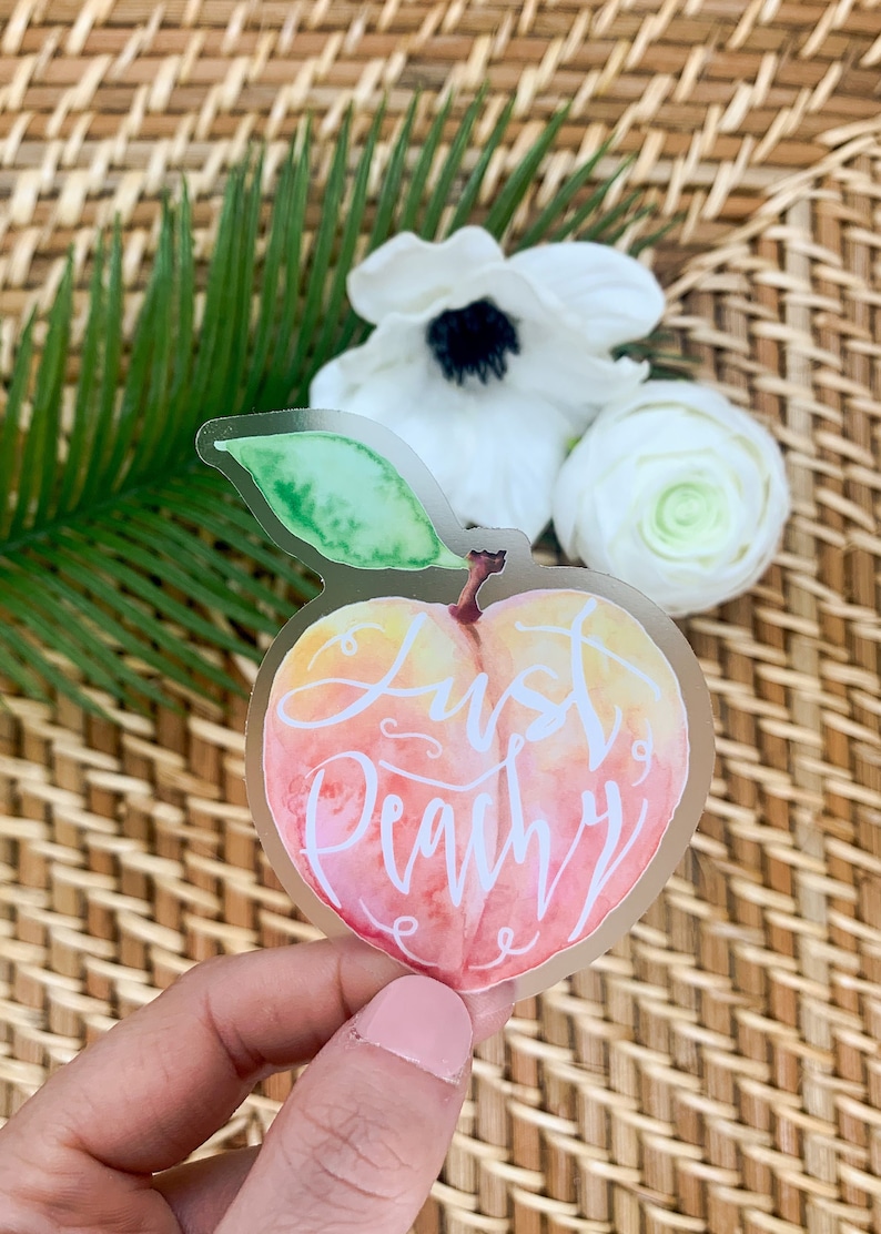 Op de afbeelding: Een transparante sticker met een aquarel perzikontwerp. De perzik is roze en oranje met een groen blad. De tekst "Just Peachy" is in wit cursief op de perzik geschreven.