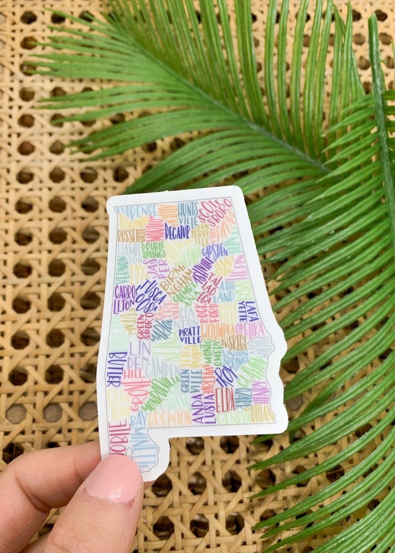 May include: A white sticker with the outline of the state of Alabama. The sticker is filled with the names of cities and towns in Alabama, written in different colors. The sticker is on a woven surface.