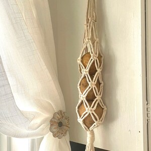 May include: A cream-colored macrame potato holder with three potatoes inside, hanging from a wooden ring. The holder is suspended against a white cabinet door. A white curtain is visible on the left.