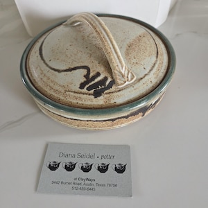 May include: A round ceramic lidded dish with a speckled beige and brown glaze, featuring a dark green rim and a curved handle on the lid. A business card for Diana Seidel potter at ClayWays 5442 Burnet Road, Austin, Texas 78756 512-459-6445 is placed in front.