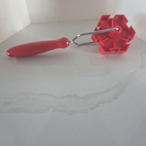 May include: A red plastic pastry roller featuring a snowflake-shaped cutting wheel attached to a metal frame and a smooth red handle. The tool is designed for creating decorative patterns in dough, with a distinct six-pointed star design.