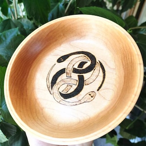 May include: A wooden bowl with a black snake design burned into the wood. The snake is curled up in a circle and has a detailed pattern of small dots.