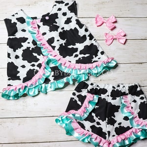 Completo per bambine con stampa mucca a balze, top in raso blu e scozzese rosa con doppio volant, pantaloncini, fiocchi, quadretti rosa e volant blu a quadretti.