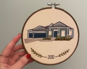 May include: Embroidered hoop art featuring a grey house with a white door and green landscaping. The hoop is decorated with green leaves and the year 2020.