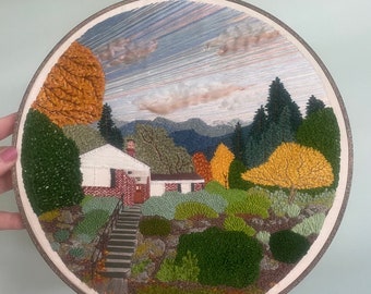 May include: Hand-stitched embroidery art in a wooden hoop. The artwork depicts a house with a red brick facade, surrounded by trees and bushes in shades of green, yellow, and orange. A mountain range is visible in the background under a cloudy sky.