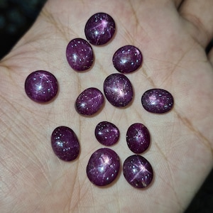 May include: A collection of ten oval-shaped, deep purple gemstones with a star-like pattern. The stones are arranged on a hand.