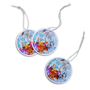 May include: Three round air fresheners with a disco ball design. Each air freshener features a mosaic of colorful squares in shades of blue, orange, yellow, and purple. They have a white border and a gray string for hanging.