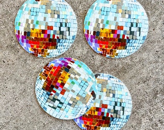 May include: Four circular coasters with a disco ball design. The coasters have a mosaic pattern of small squares in shades of blue, silver, red, orange, and purple. They are arranged on a grey concrete surface.